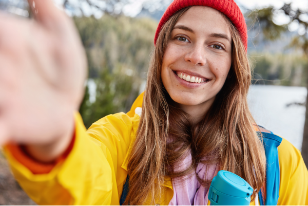 optimistic-female-model-has-toothy-smile-stretches-hand-as-makes-selfie-dressed-casually